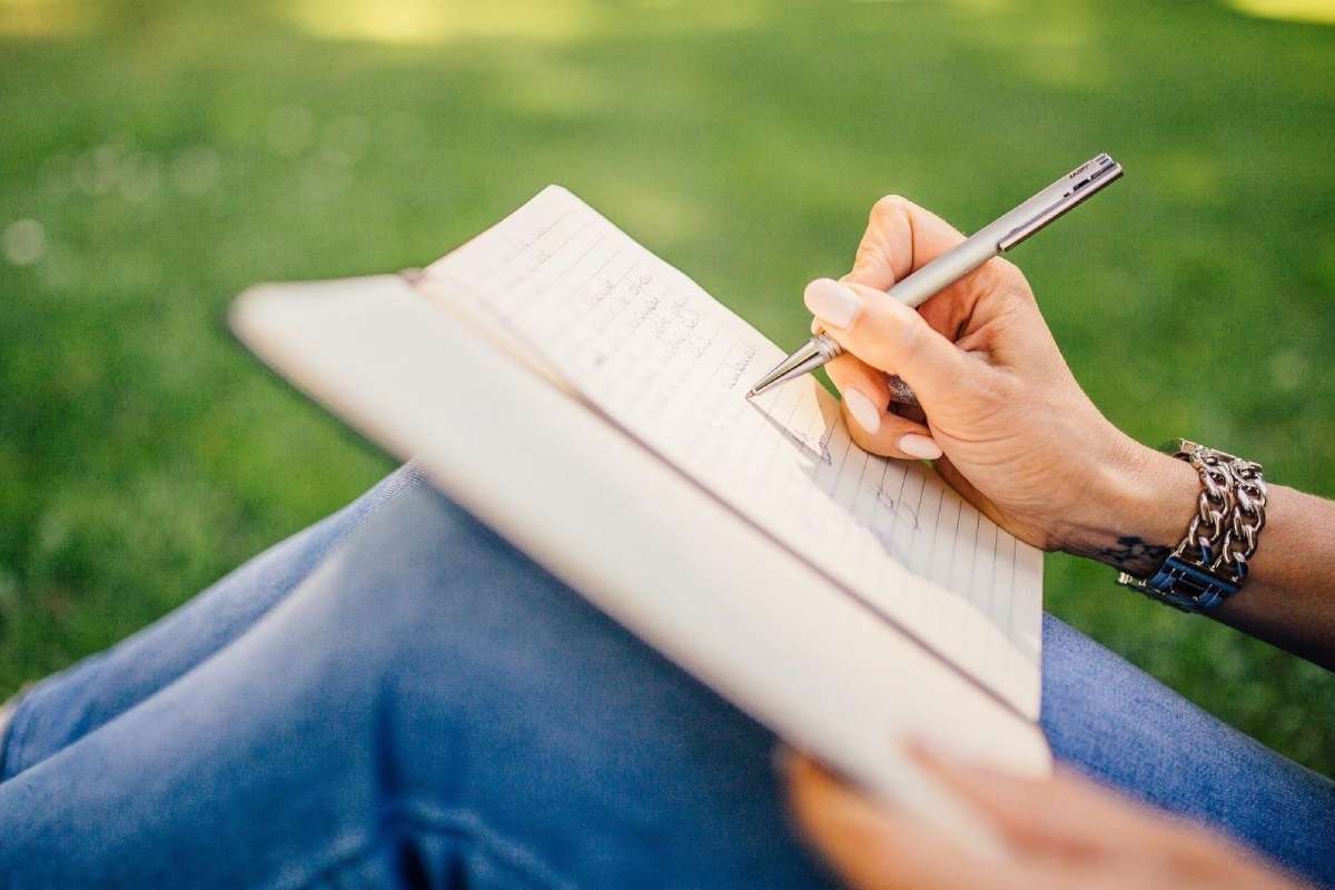 GACL award winners 2025 1 - TA NEA Person writes in an open notebook while sitting on green grass, outdoors, with a silver pen and bracelets on the wrist.